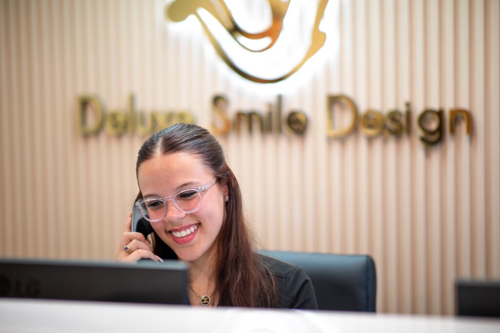 Woman smiling while talking on the phone at a reception desk with "Deluxe Smile Design" sign in the background.